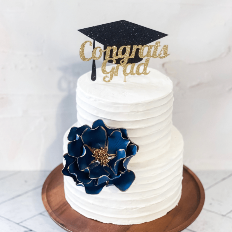 Extra large navy and gold edged open rose rose sugar flower decorating a two tier buttercream cake with a cake topper that says congrats grad