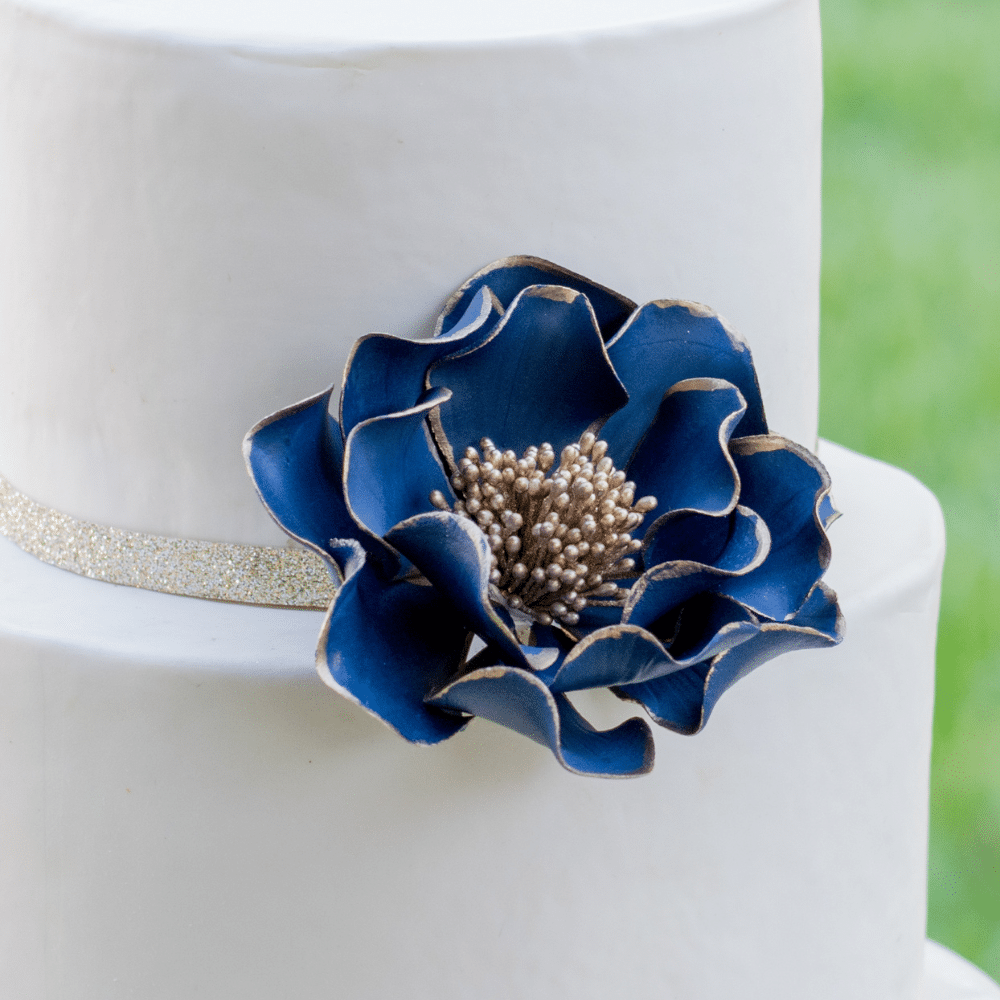 Close up of navy and gold edged open rose gumpaste flower decorating a white fondant cake
