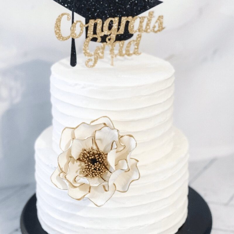 white and gold edged open rose sugar flower on a two tier buttercream cake with a graduation cake topper