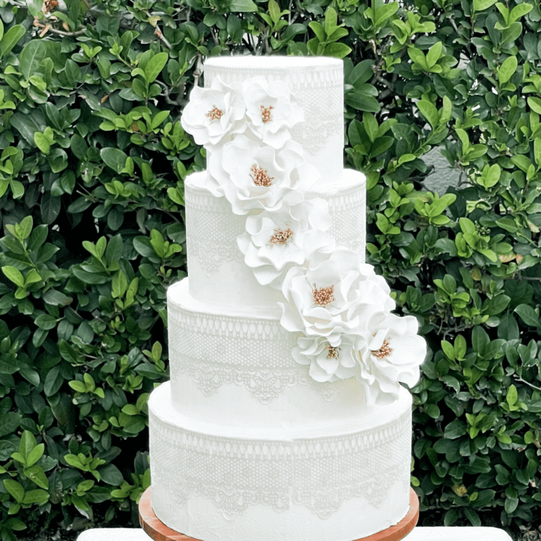four-tiered white wedding cake with intricate lace designs and a cascade of white and gold open rose sugar flowers, displayed on a wooden cake stand against a background of greenery