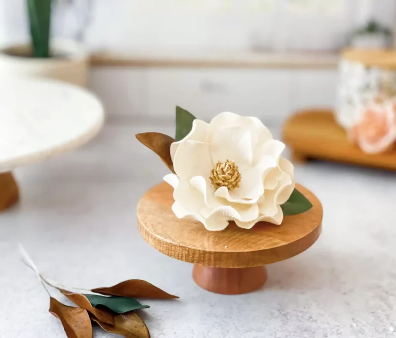magnolia sugar flower in white and gold with brown and green leaves displayed on a wooden cake stand