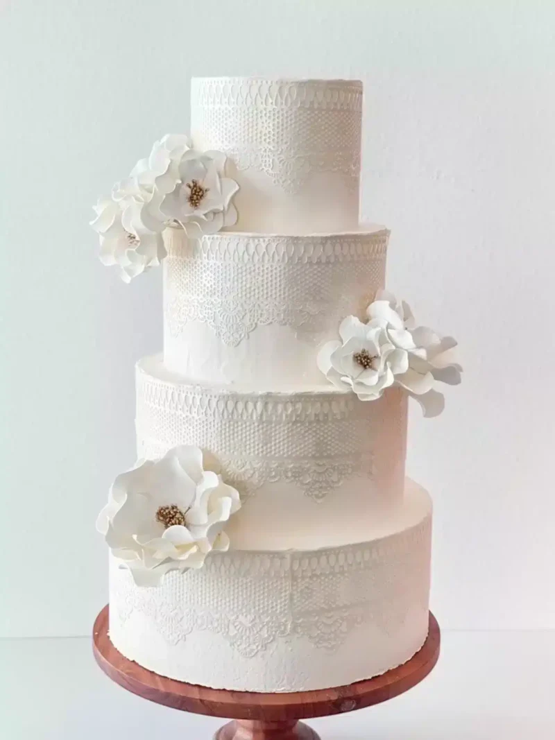 four-tiered white wedding cake with intricate lace designs and large white and gold open rose sugar flowers, displayed on a wooden cake stand.