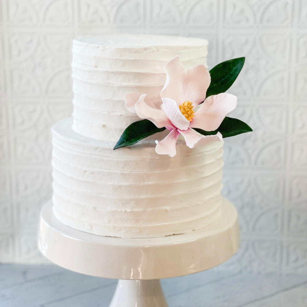 pink saucer magnolia sugar flower on a two tier white fondant buttercream cake