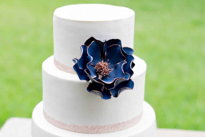 Extra large navy and rose gold edged open rose sugar flower displayed on a two tier white fondant cake