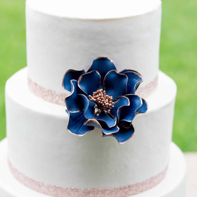 Close up of small navy and rose gold edged open rose sugar flower on a white fondant cake