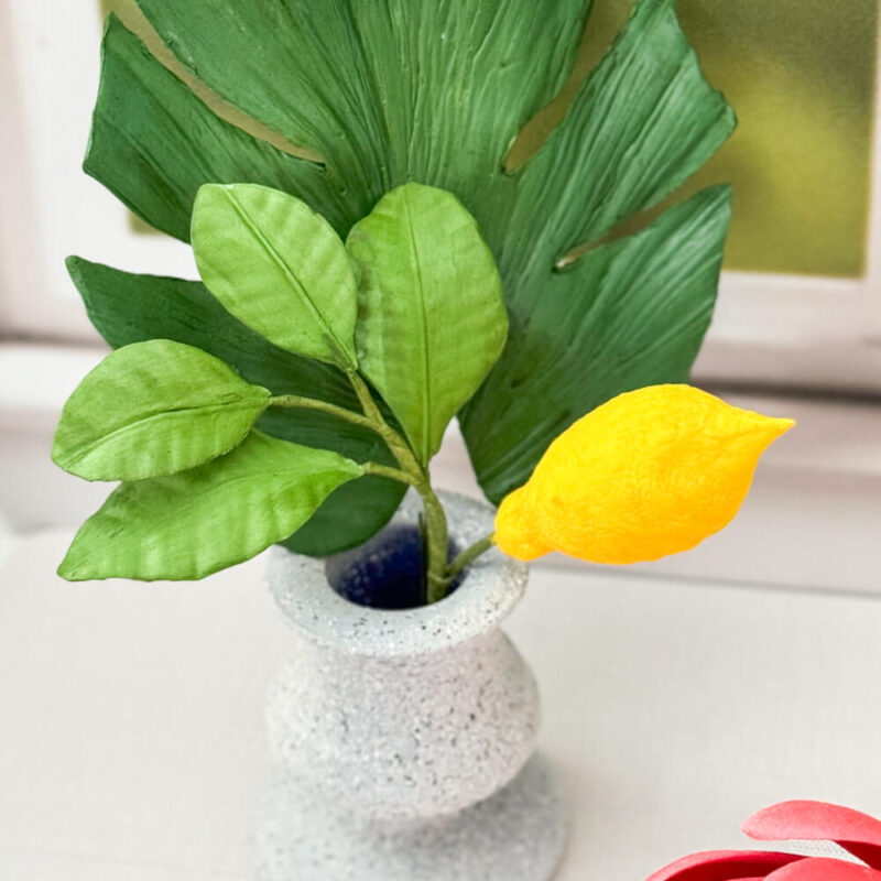 A gumpaste vibrant yellow lemon, paired with three realistic green leaves, displayed in a gray textured vase.