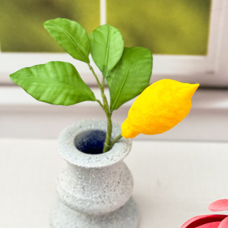 A gumpaste vibrant yellow lemon, paired with three realistic green leaves, displayed in a gray textured vase.