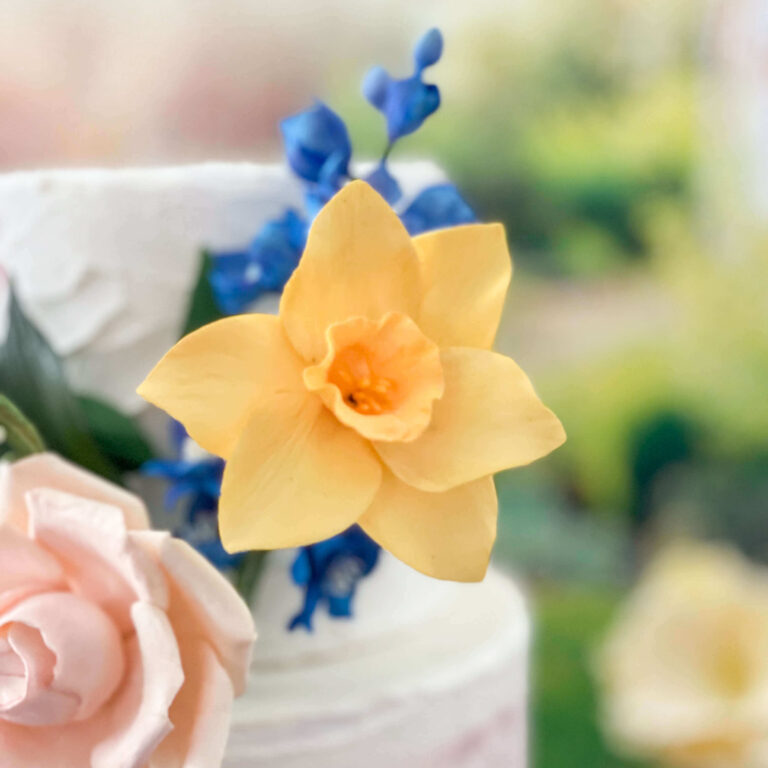 Close-up of a yellow daffodil and a pink rose sugar flower on a white iced cake, with a blurred background of greenery and blue flowers.