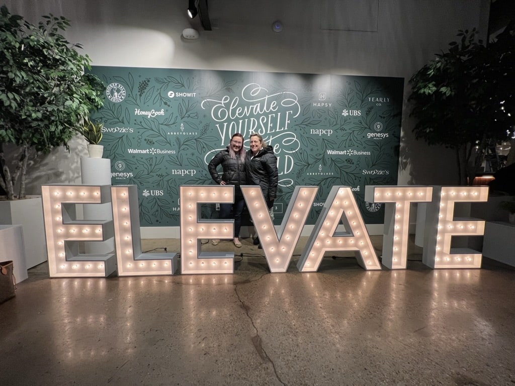 photo of two women standing behind a lighted sign that says ELEVATE