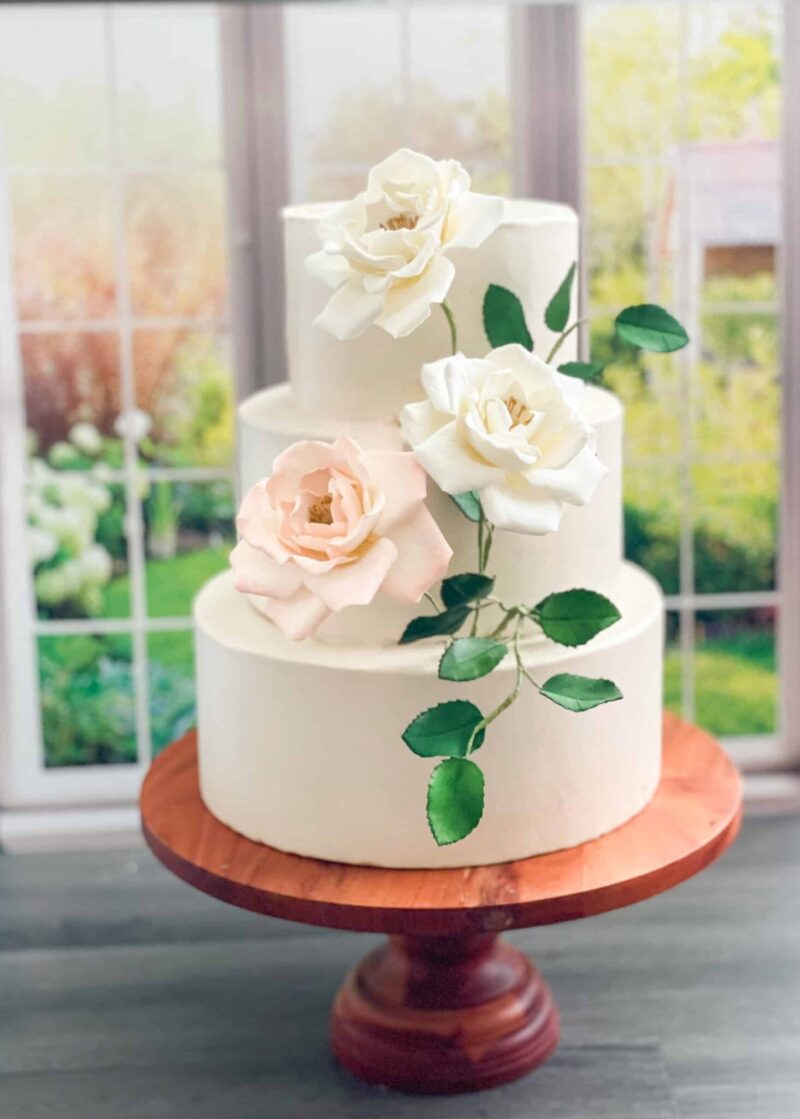 Three tier white wedding cake featuring sugar flower cream and blush roses and green leaves cascading down the cake