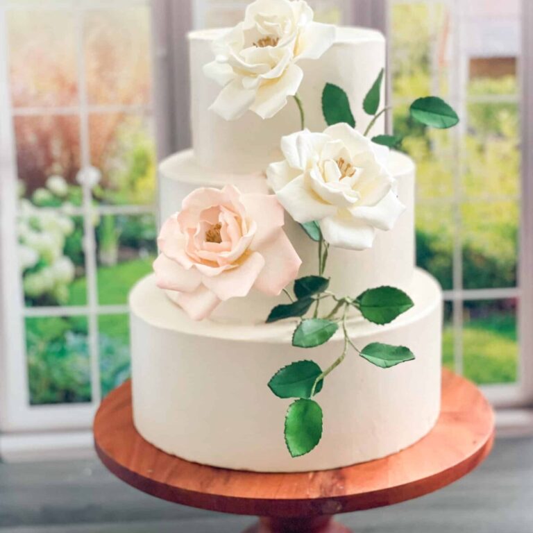 Three tier white wedding cake featuring sugar flower cream and blush roses and green leaves cascading down the cake