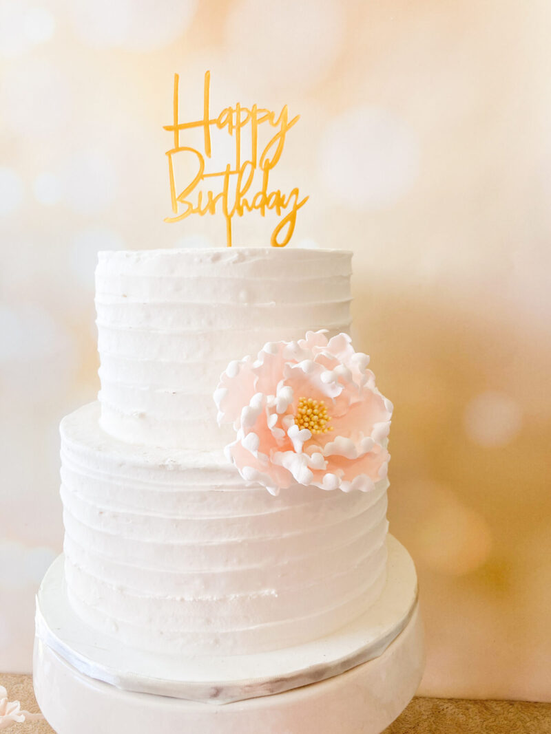 blush and yellow peony sugar flower On a two tier textured buttercream cake with a gold “happy birthday” acrylic cake topper on top