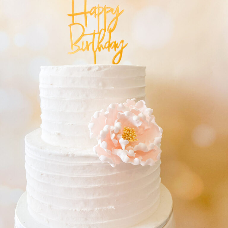 blush and yellow peony sugar flower On a two tier textured buttercream cake with a gold “happy birthday” acrylic cake topper on top