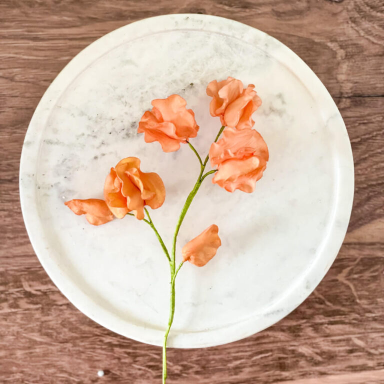 A small stem of coral colored sweet pea sugar flowers rests on a circular white marble cake stand, set against a wooden background.