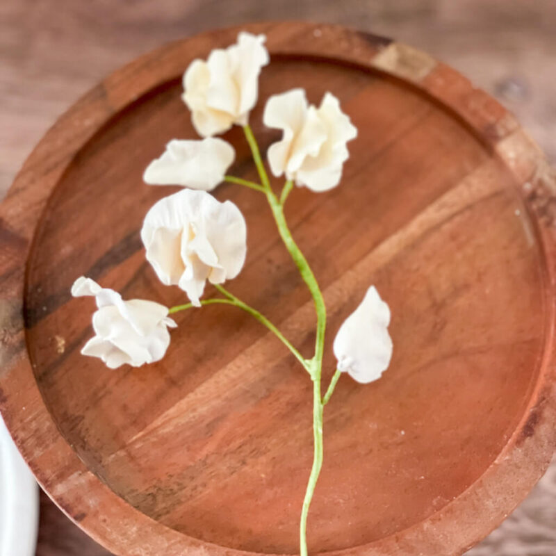 A small stem of cream colored sweet pea sugar flowers rests on a circular white marble cake stand, set against a wooden background.