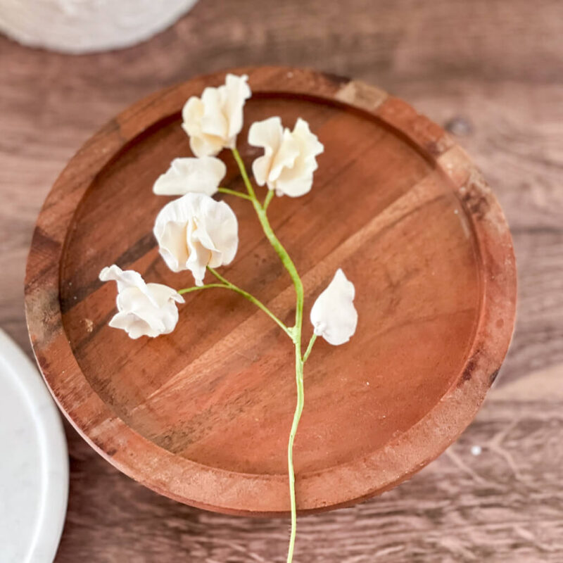 A small stem of ivory colored sweet pea sugar flowers rests on a circular white marble cake stand, set against a wooden background.