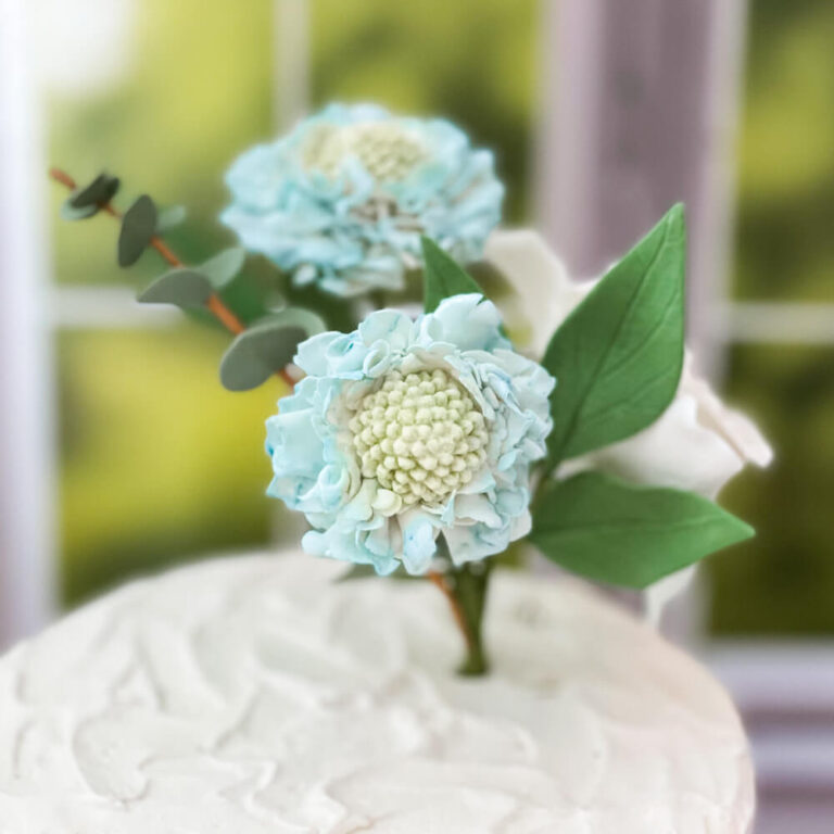 blue scabiosa sugar flower surrounded by leaves and placed in a buttercream cake