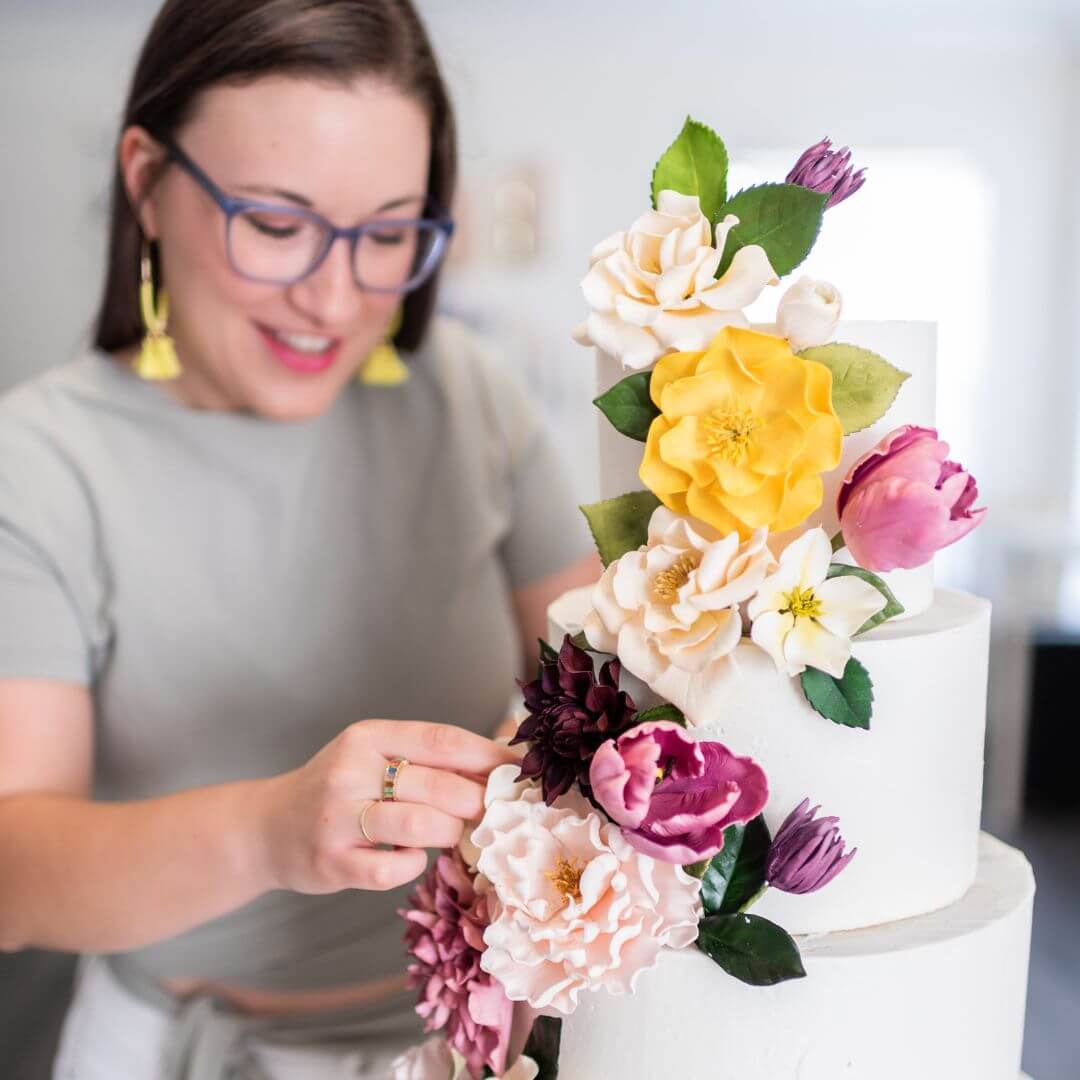 woman putting sugar flowers on a three tier wedding cake cascing with pink and purple sugar flowers